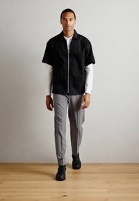 Cămașă neagră cu mânecă scurtă, cu fermoar, purtată peste un top alb cu mânecă lungă, asortată cu pantaloni gri slim-fit și adidași negri, pe un podea din lemn.
