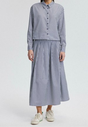 Personne portant une chemise à rayures verticales bleues et blanches avec boutons et une jupe large plissée assortie, avec des baskets blanches sur fond uni.