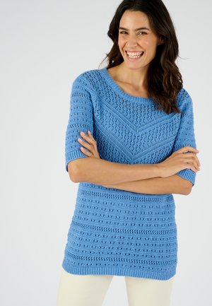 Pull en tricot bleuâtre à manches courtes, avec un motif texturé, des poignets côtelés et un large col. Associé à un pantalon de couleur claire.