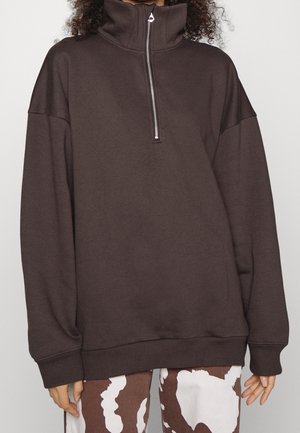 Personne portant un pull ample marron foncé avec col demi-fermeture éclair et pantalon marron et blanc à motif, se tenant devant un fond uni.