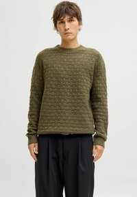 Person with short brown hair wearing an olive green textured knit sweater and loose black pants, standing against a plain light background.