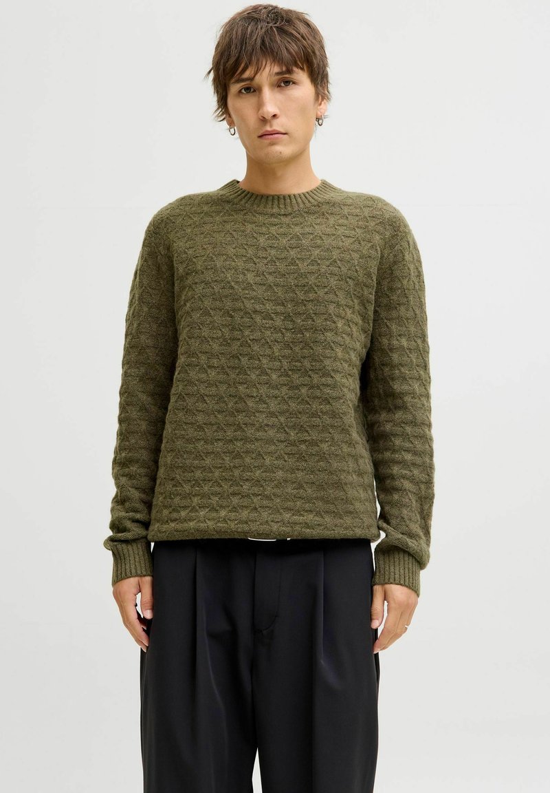 Person with short brown hair wearing an olive green textured knit sweater and loose black pants, standing against a plain light background.