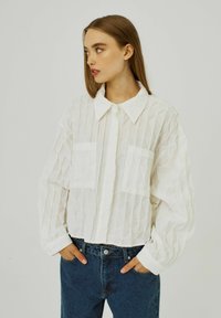 Vit, texturerad skjorta med knäppning fram, spetsig krage och två bröstfickor, tillsammans med blå högmidjade jeans.