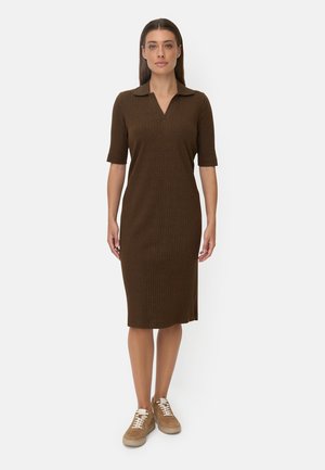 Frau, die nach vorne schaut und ein braunes, knielanges, geripptes Kleid mit ellbogenlangen Ärmeln sowie beige Sneaker trägt.