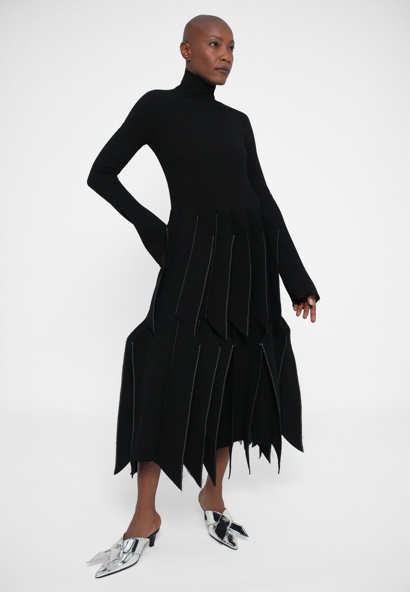 Black dress with long sleeves, high neck, and layered asymmetric hem. Features silver stitching; paired with silver heeled mules.