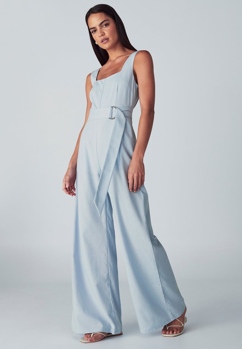 Ženska, oblečena v svetlo modro brezrokavnik širokega kroja z mestom za pas in kvadratnim dekoltejem, nosi sandale z odprtim prstom na enobarvni podlagi.