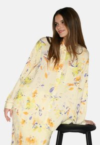 Blusa gialla fluida con maniche lunghe e motivo floreale in viola e arancione. Presenta un'apertura con bottoni davanti e una singola tasca sul petto.