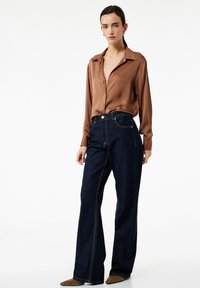 Camisa marrón de botones y mangas largas combinada con unos jeans acampanados de denim azul oscuro de tiro alto, que presentan costuras en contraste y bolsillos.