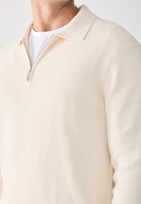 Suéter beige con cremallera y textura acanalada, con diseño de cuello. Presenta una cremallera plateada y un aspecto de capas con una camiseta blanca debajo.