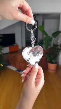 Silberner herzförmiger Schlüsselanhänger mit der Gravur "tommy jeans", befestigt an einer Metallkette und einem Schlüsselring, gehalten vor einem unscharfen Hintergrund.