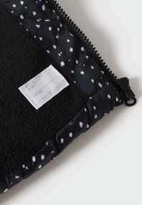 Černá zipová taštička s texturovaným fleece povrchem a podšívkou s puntíky. Obsahuje bílou látkovou etiketu na jméno a telefonní údaje.