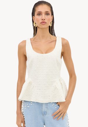 Femme aux cheveux mouillés tirés en arrière portant un top blanc sans manches à basque orné de perles et un jean bleu clair avec des détails en perles, posant avec la main dans la poche.
