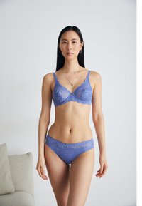 Lila spetsbehå och matchande trosor har vågiga kanter, intrikata blommönster och justerbara remmar. Modellen står i en neutral miljö.