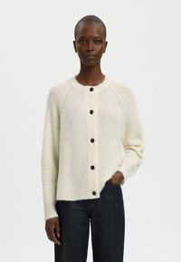 Krems balts džemperis, kas izgatavots no mīkstas, faktūrētas auduma. Iezīmējās ar apaļu kaklu, septiņām tumšām pogām un raglāna piedurknēm. Valkāts ar tumšiem džinsiem.