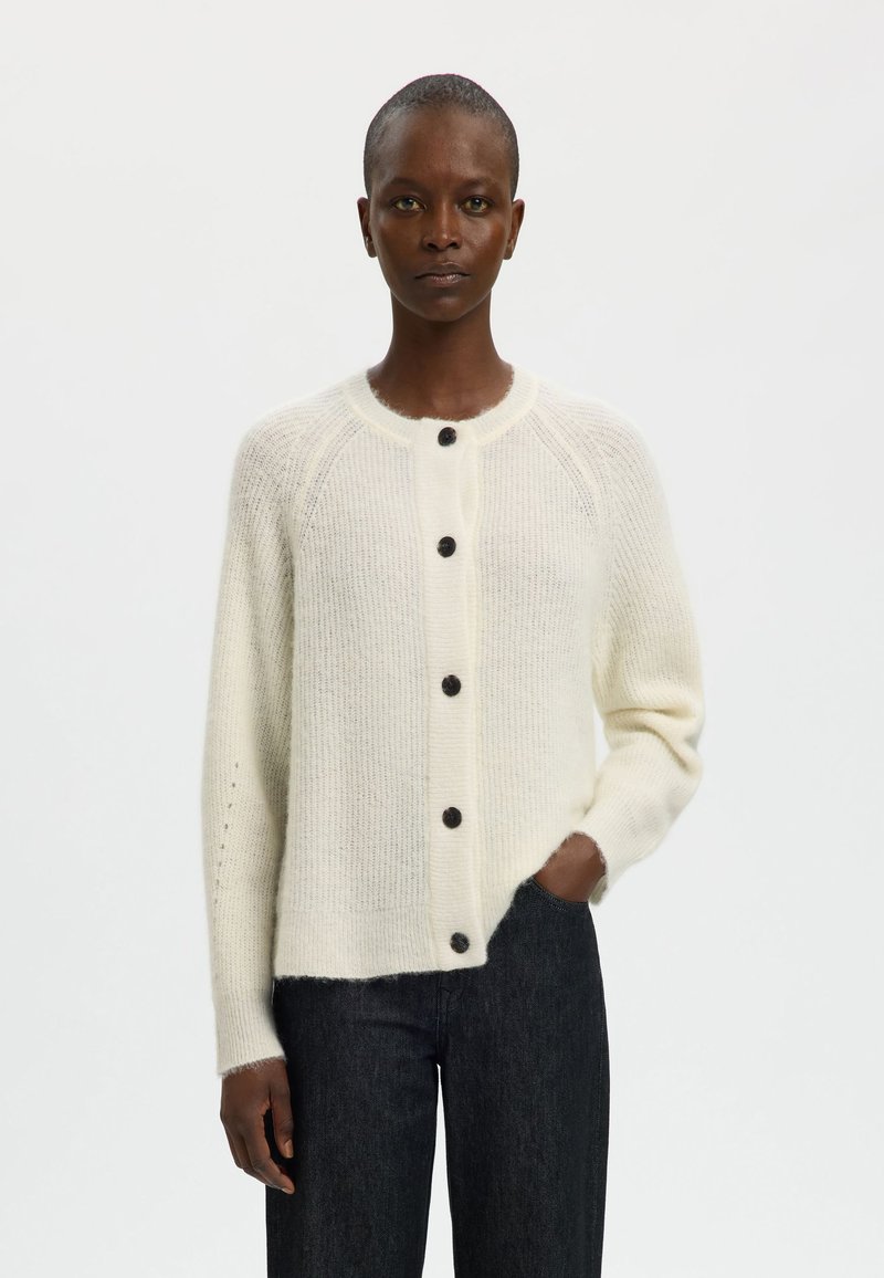 Krems balts džemperis, kas izgatavots no mīkstas, faktūrētas auduma. Iezīmējās ar apaļu kaklu, septiņām tumšām pogām un raglāna piedurknēm. Valkāts ar tumšiem džinsiem.