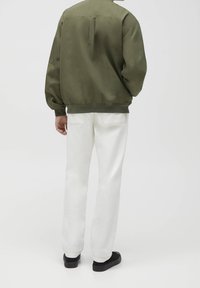 Kurtka typu bomber w oliwkowo-zielonym kolorze o luźnym kroju, zestawiona z białymi spodniami o prostych nogawkach i czarnymi butami, pokazana od tyłu.