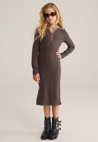 Gestreiftes Langarmkleid in Braun und Beige mit Henley-Ausschnitt und Knöpfen. Kombiniert mit schwarzen Stiefeletten mit drei verstellbaren Riemen.