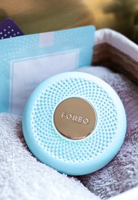 Blå silikon ansiktsrenseapparat med teksturert overflate og gull-logo. Plassert på et hvitt håndkle, sammen med en blå pakke.