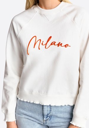 Sweat-shirt blanc avec ourlet festonné et poignets, texte orange « Milano » sur la poitrine, porté avec un jean en denim bleu clair.