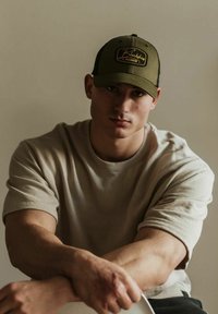Barna T-shirt és olíva zöld snapback sapka logóval. A személynek rövid haja van, ül és karját térdére támasztja. Semleges háttér.
