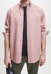 Camicetta button-up rosa chiaro con colletto, maniche lunghe e bottoni bianchi, indossata sopra una maglietta nera, abbinata a pantaloni verde scuro.