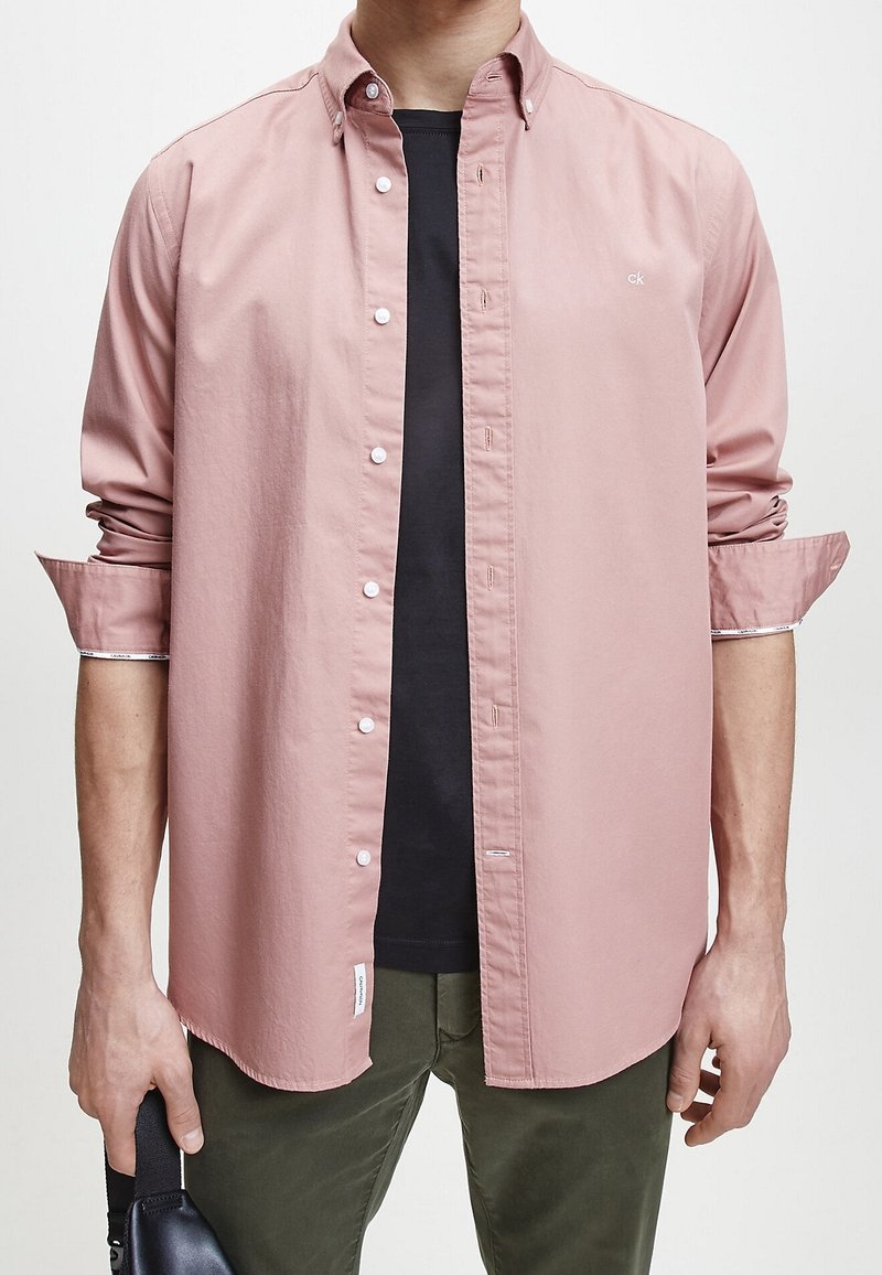Camicetta button-up rosa chiaro con colletto, maniche lunghe e bottoni bianchi, indossata sopra una maglietta nera, abbinata a pantaloni verde scuro.