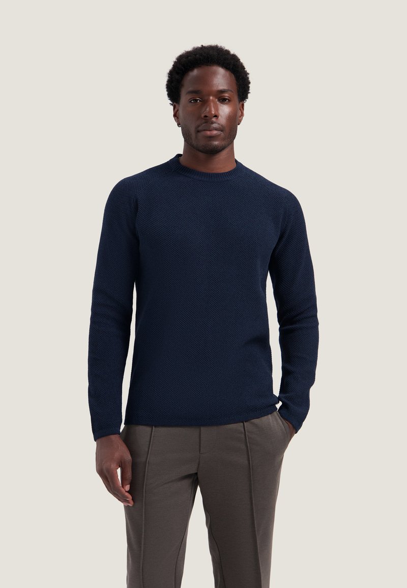 Homme debout, les mains partiellement dans les poches, portant un pull texturé bleu marine et un pantalon gris sur un fond clair uni.