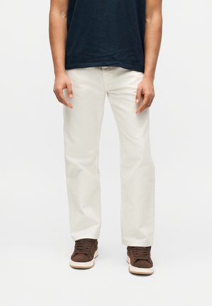 Person trägt cremefarbene, gerade geschnittene Hose, dunkelbraune Sneakers mit weißen Sohlen und ein schwarzes Kurzarmshirt, steht vor einem einfarbigen Hintergrund.