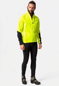 Jachetă softshell galbenă strălucitoare, cu mâneci negre, buzunare cu fermoar, guler înalt, asortată cu pantaloni negri mulați și încălțăminte neagră robustă.