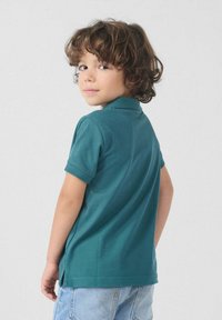 Jong kind met krullend bruin haar, gekleed in een tealgroene polo en lichtblauwe spijkerbroek, kijkt over de schouder tegen een effen achtergrond.