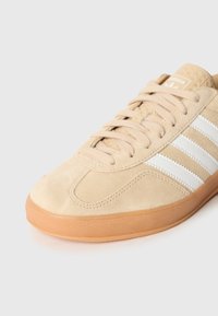 Bēšas zamšādas kedas ar trim baltām svītrām, teksturētām šņorēm un gumijas zoli. Apaļa purngala forma ar izšūtiem detaļām.