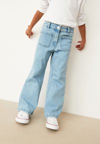 Jeans din denim de culoare albastru deschis, cu un design cu picioare largi, stil cu cinci buzunare și sistem de închidere clasic cu nasture și fermoar. Asortați cu adidași albi.