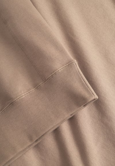 Tessuto di cotone marrone chiaro caratterizzato da una texture liscia, bordi cuciti e un dettaglio a risvolto. Il materiale appare morbido e leggero.