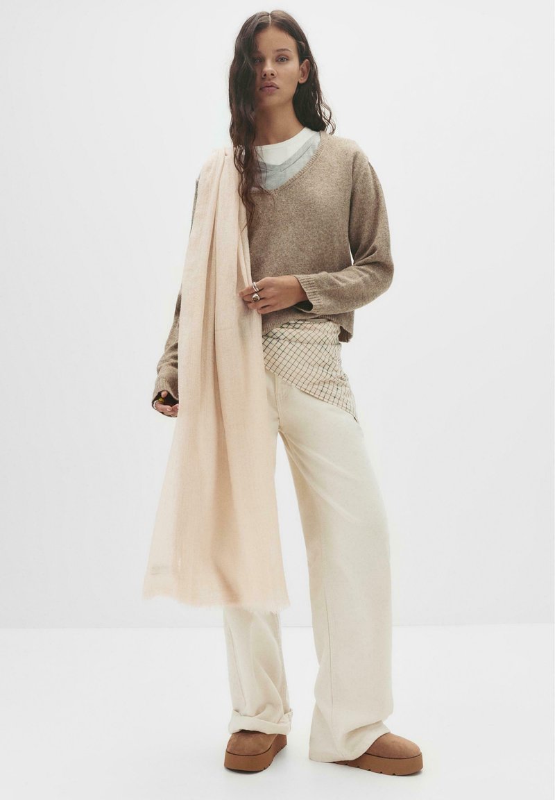 Maglione marrone a maglia sopra una camicia bianca; pantaloni larghi crema con sovrapposizione a motivi; morbida sciarpa rosa drappeggiata; scarpe a piattaforma marroni.