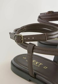 Tmavohnedá kožená sandála s nastaviteľným remienkom so zlatou mosadznou prackou a čiernou podrážkou, zobrazená v detailnom zábere.