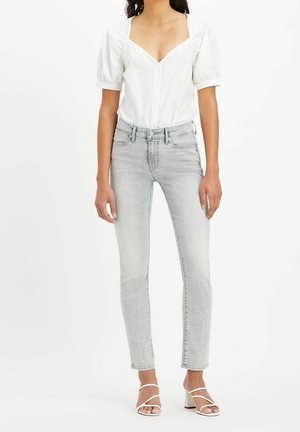 Femme portant un chemisier blanc à manches bouffantes, un jean skinny taille haute gris clair et des sandales blanches à brides avec talons carrés, debout sur fond blanc.