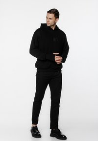 Czarna bluza z kapturem z kieszenią z przodu, miękka w dotyku, z małą naszywką z logo. Połączona z czarnymi spodniami slim-fit i błyszczącymi czarnymi butami.