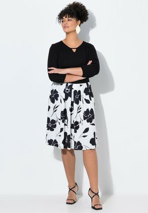 Schwarzes Langarmoberteil mit einem Schlüssellochdetail, kombiniert mit einem weißen Faltenrock mit schwarzen Blumenmustern. Schwarze Riemchensandalen runden den Look ab.