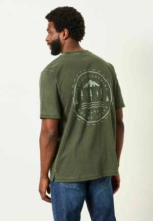 Mann trägt ein olivgrünes T-Shirt mit einem Outdoor-Themen-Grafik und Text auf dem Rücken, kombiniert mit blauen Jeans.