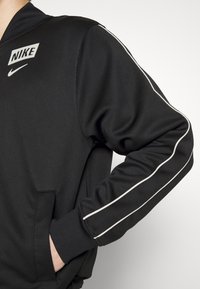 Felpa Nike nera con zip frontale, caratterizzata da un colletto, tasca laterale e strisce bianche sui lati delle maniche. Tessuto morbido.