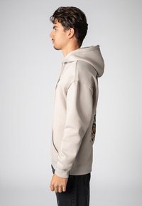 Beige hoodie met een voorzak in kangoeroestijl en een grafisch ontwerp. De stof voelt zacht aan, heeft een ontspannen pasvorm en een verstelbare capuchon met trekkoord.