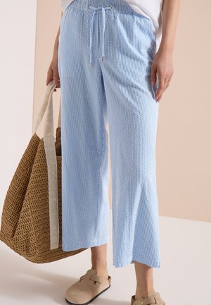 Femme portant un pantalon large à rayures bleu clair et blanc, des chaussures beiges sans lacets, et tenant un grand sac cabas tissé marron.