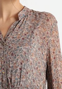 Blouse à motifs floraux en tissu léger et transparent. Dotée d'un col montant, d'une fermeture à boutons et de couleurs douces comme le beige, le bleu, l'orange et le bordeaux.