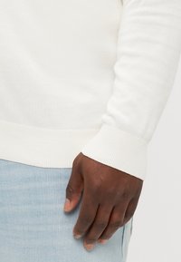 Fehér, bordázott kötött pulóver, fitted mandzsettákkal, egy személy karján bemutatva, világoskék farmernadrággal párosítva. Simaság jellemzi, klasszikus dizájn.