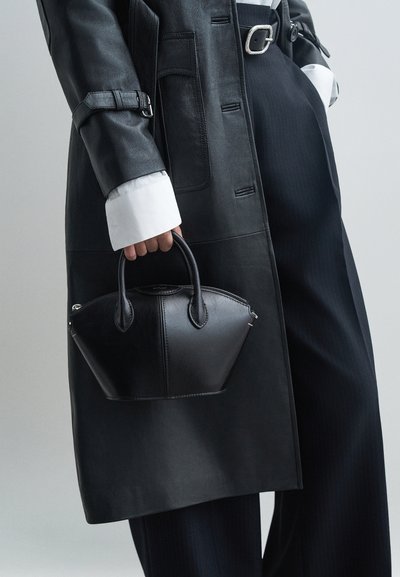 Sac à main en cuir noir avec une forme structurée, doté de poignées courtes, de coutures discrètes et d'accents en métal argenté, tenu contre une tenue sombre.