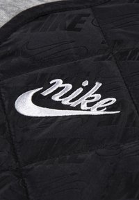 Tissu matelassé noir arborant un logo Nike brodé en blanc, avec des motifs subtils en noir sur noir en arrière-plan. La texture semble lisse.