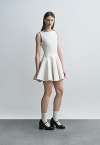 Joven mujer con un vestido blanco sin mangas y texturizado, calcetines beige y zapatos Mary Jane de plataforma negros, posando frente a un fondo gris liso.