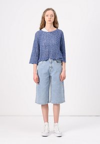 Pull en tricot bleu à manches trois-quarts associé à des culottes en denim bleu clair taille haute et des baskets blanches, se tenant sur un fond uni.