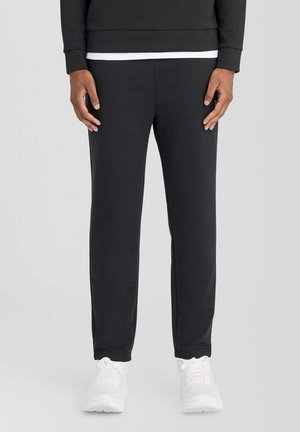 Person iført sorte joggingbukser, sort langærmet top og hvide sneakers, stående foran en ensfarvet lys baggrund.