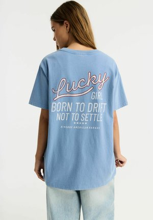 Młoda kobieta z długimi włosami nosi luźny niebieski T-shirt z napisem "Lucky girl born to drift not to settle" na plecach.